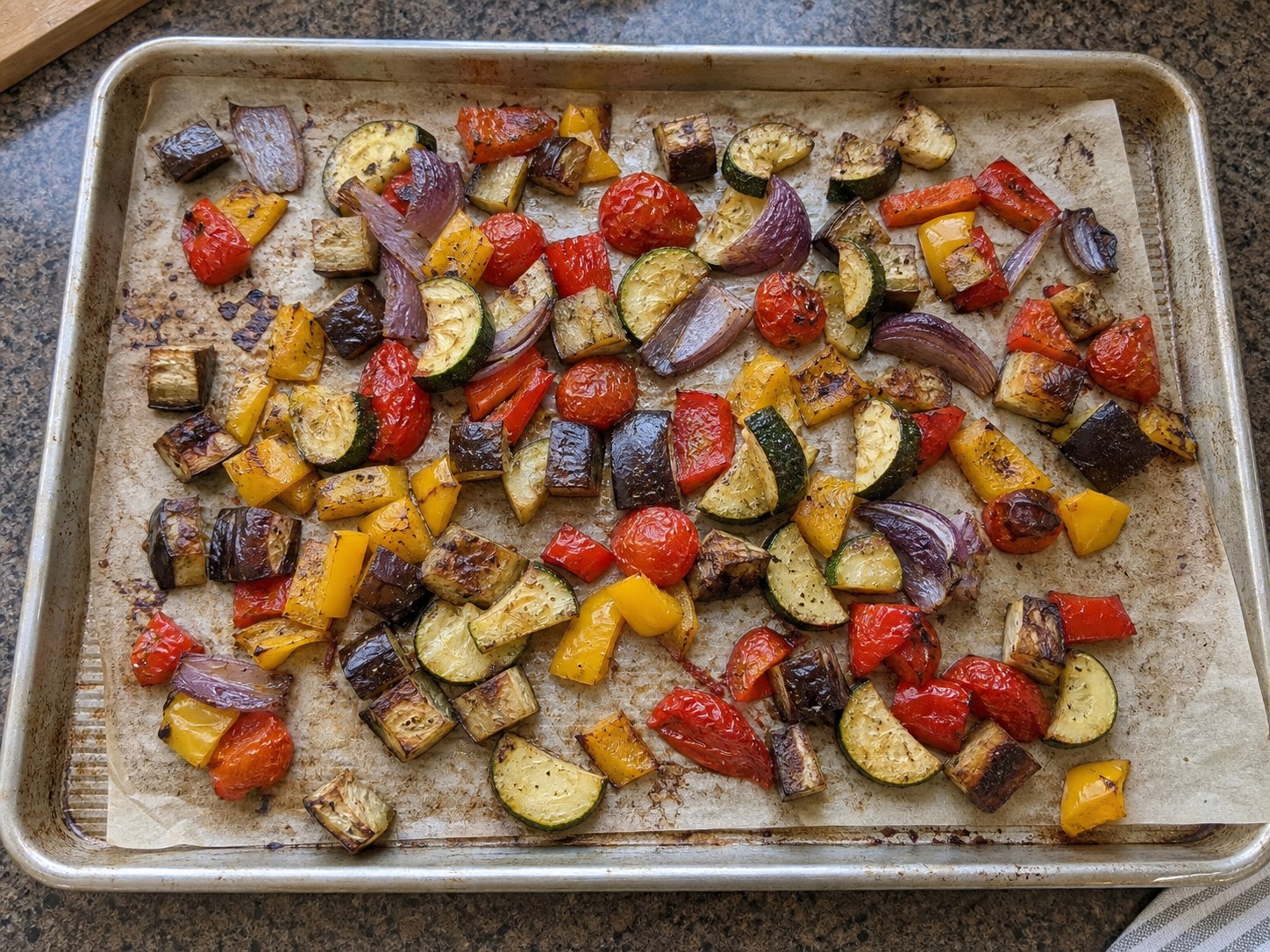 Roasted eggplant, zucchini, bell peppers, red onion, and cherry tomatoes cooling on a sheet pan for couscous salad.