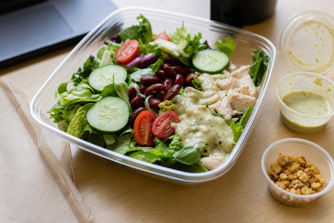 An open lunch salad with dressing partly poured and dry crunchy topping still in a separate cup.