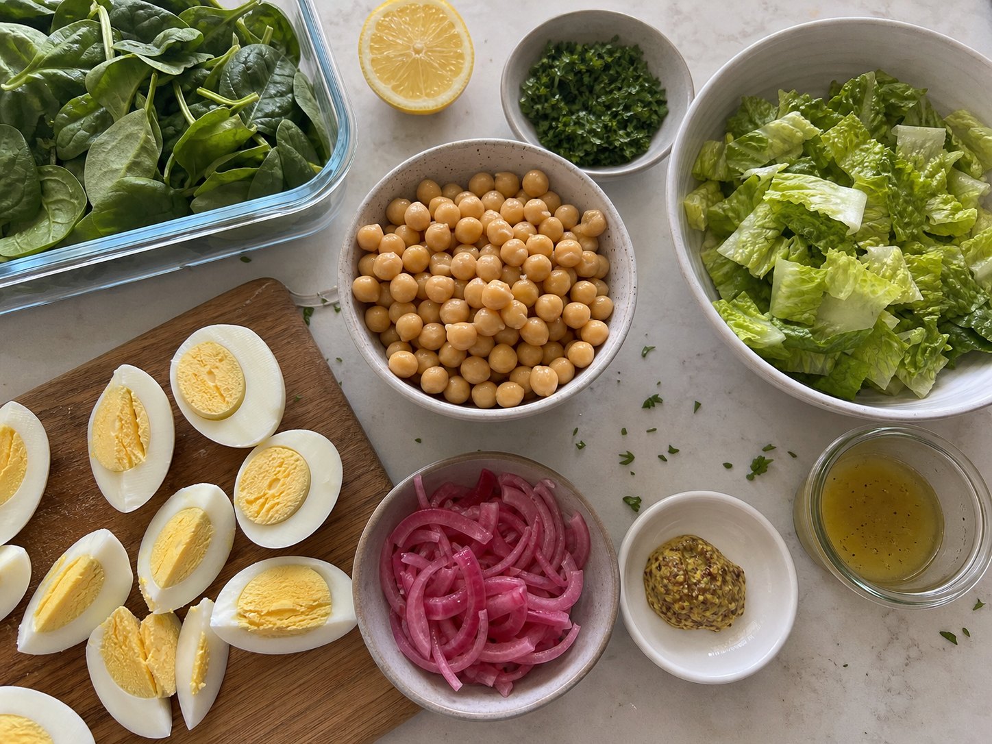 Egg and chickpea protein salad ingredients with hard-boiled eggs, chickpeas, spinach, romaine, pickled red onions, herbs, lemon, mustard, and dressing cup.