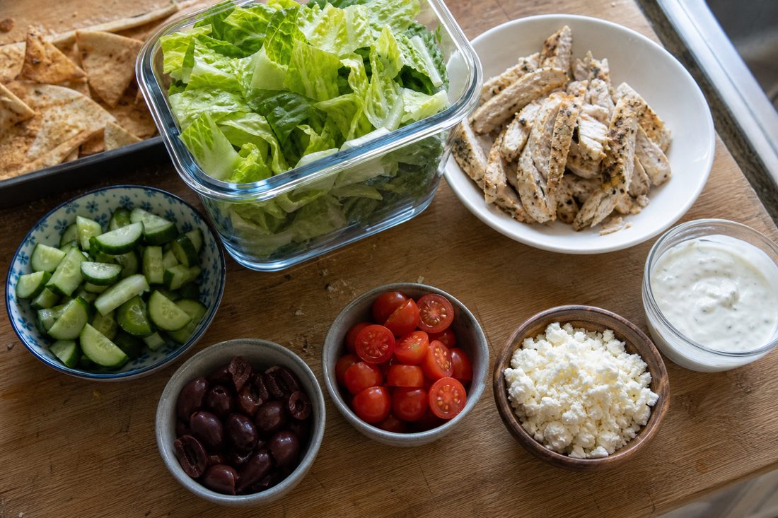 Greek chicken salad lunch box ingredients with lemon chicken, romaine, cucumber, tomatoes, olives, feta, yogurt dressing, and pita chips.