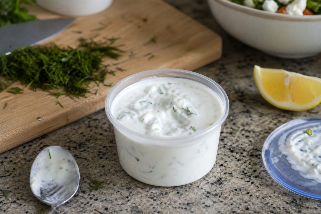 Small cup of garlic yogurt dressing with herbs and lemon beside a Greek chicken salad setup.