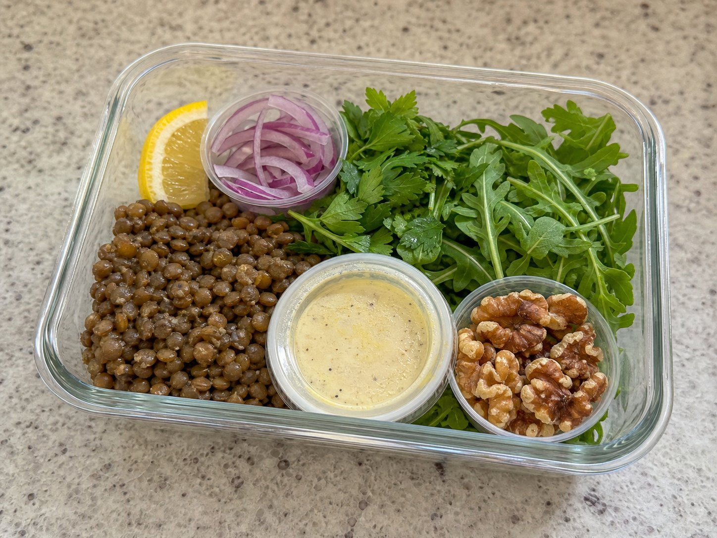 Unmixed lentil lemon Dijon salad packed in a glass lunch container with lentils, arugula, parsley, red onion, dressing cup, walnuts, and lemon.
