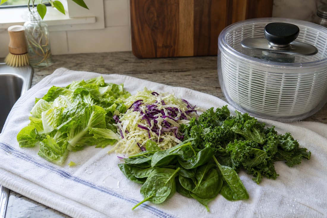 Washed romaine, cabbage, kale, and spinach drying on a towel beside a salad spinner.