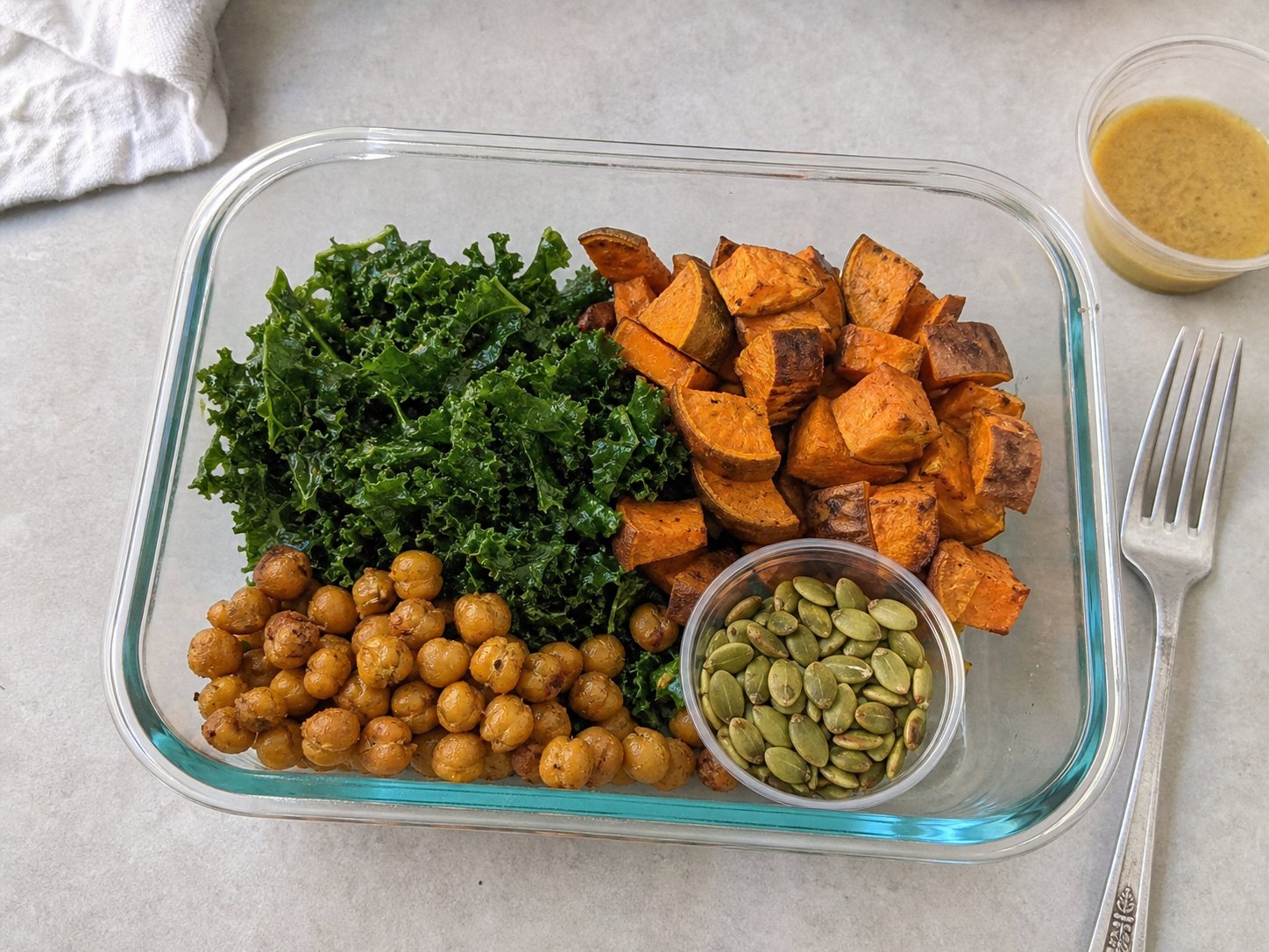Unmixed roasted sweet potato kale salad packed in a glass lunch container with kale, roasted sweet potatoes, chickpeas, maple Dijon dressing, and pumpkin seeds.